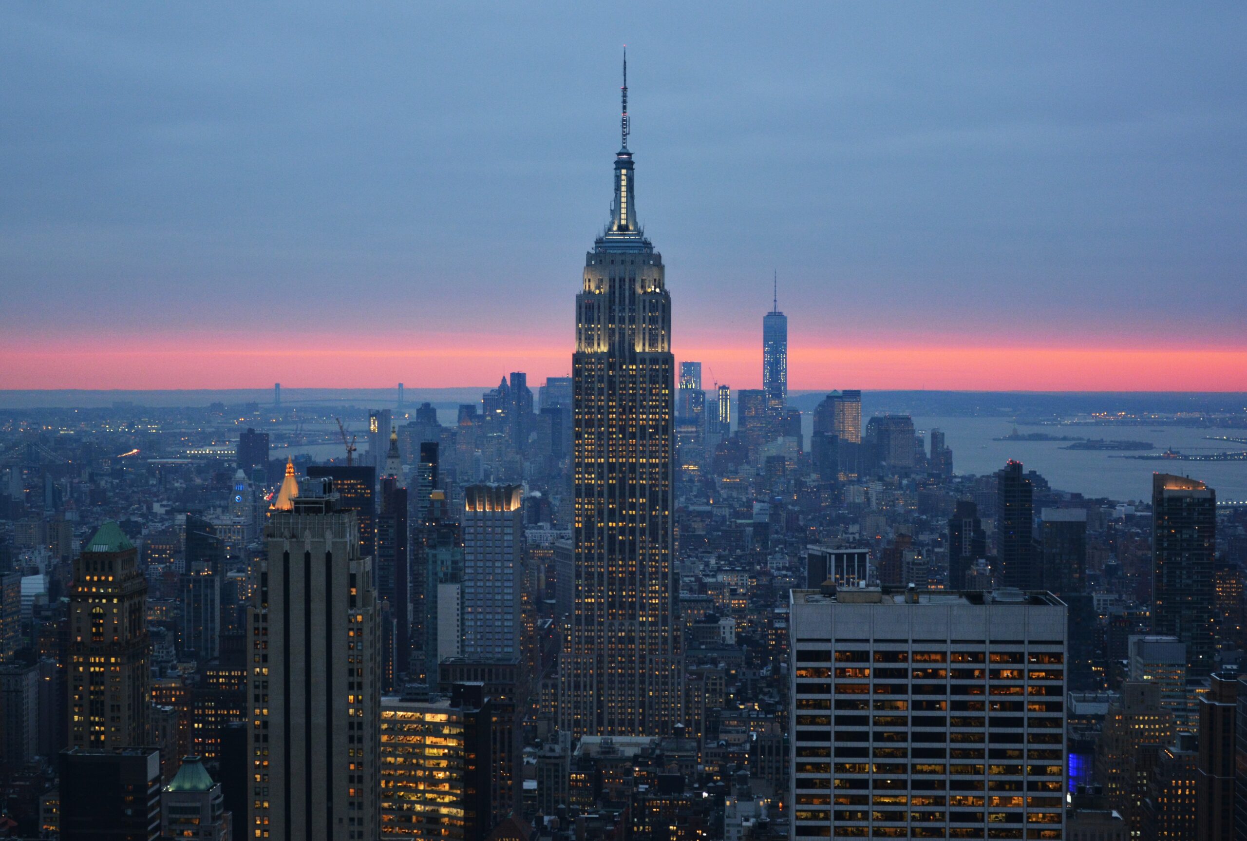 Vista panorámica del horizonte de la ciudad de Nueva York al atardecer. El Empire State Building se destaca en el centro, iluminado contra el cielo de tonos rosados y azules. A su alrededor, se ven numerosos rascacielos con luces encendidas, bajo un cielo que cambia de azul a naranja y rosa, reflejando el atardecer sobre la ciudad y el río al fondo.